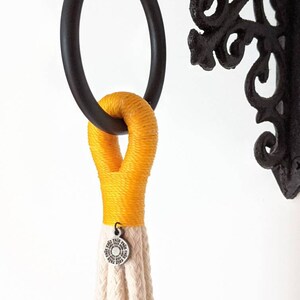 May include: A yellow macrame plant hanger with a white braided cord and a silver metal ring. The hanger is attached to a black metal hook.