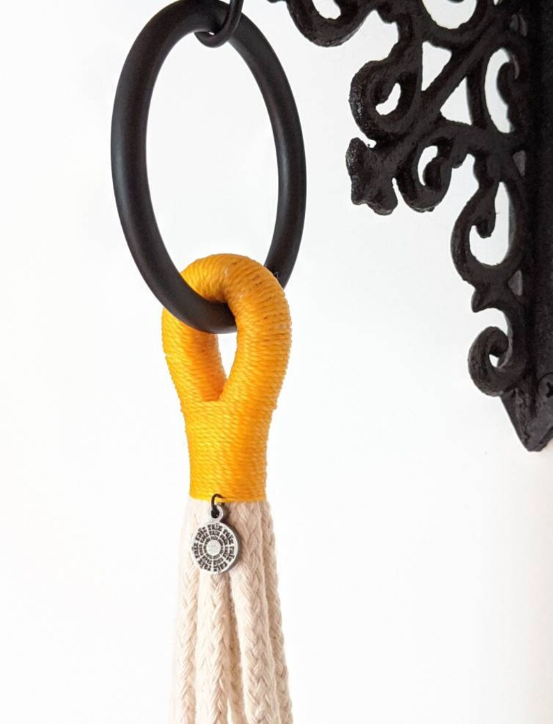 May include: A yellow macrame plant hanger with a white braided cord and a silver metal ring. The hanger is attached to a black metal hook.