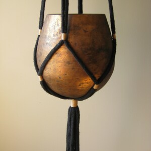 May include: A black macrame plant hanger with a copper-colored pot. The hanger has a tassel at the bottom.
