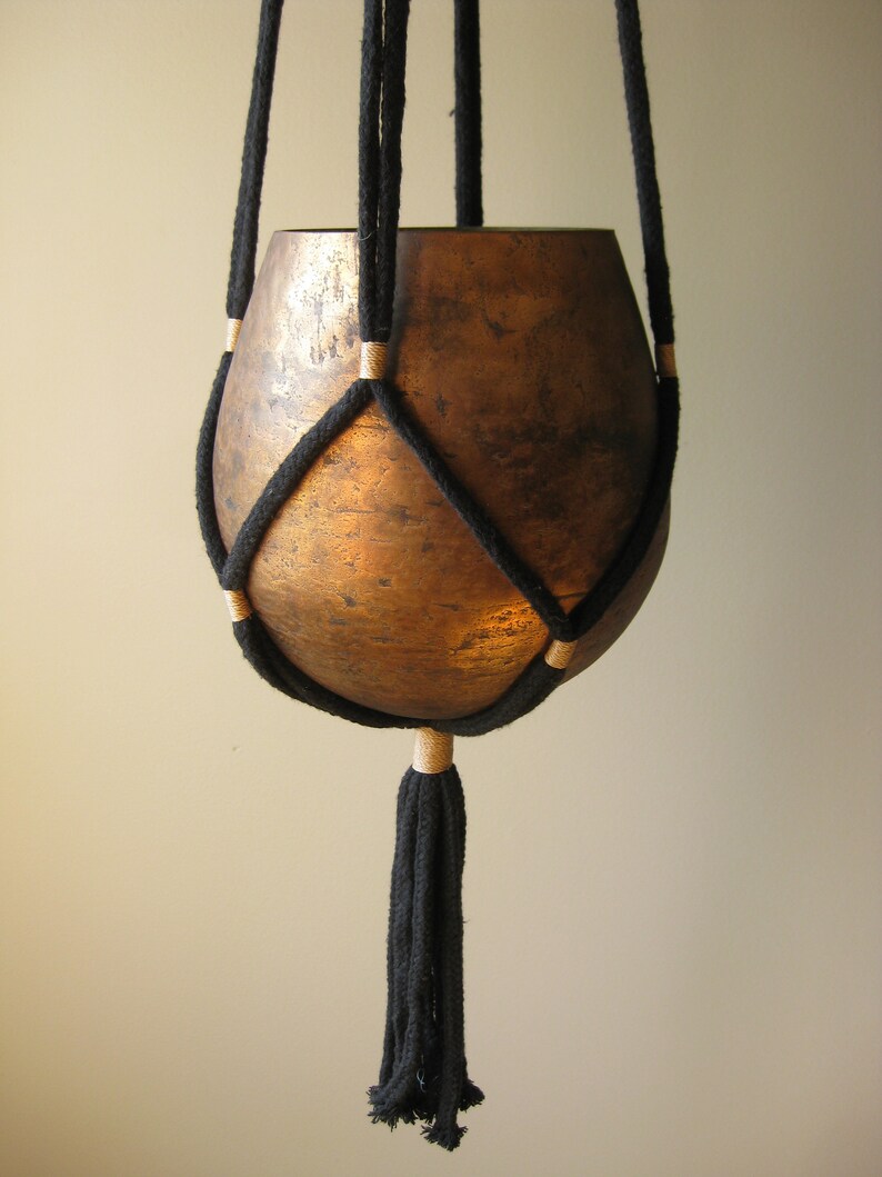 May include: A black macrame plant hanger with a copper-colored pot. The hanger has a tassel at the bottom.