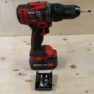Pode incluir: Uma furadeira sem fio vermelha e preta com uma bateria de 20V e uma capacidade de 3Ah. A furadeira está sobre uma superfície de madeira com um pequeno recipiente preto de parafusos.