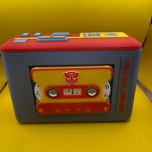 May include: A rectangular grey, red, and yellow plastic cassette player case featuring a yellow cassette tape with a red Autobot symbol in the centre. The side of the case displays the text TILL ALL ARE ONE in red letters against a grey background.