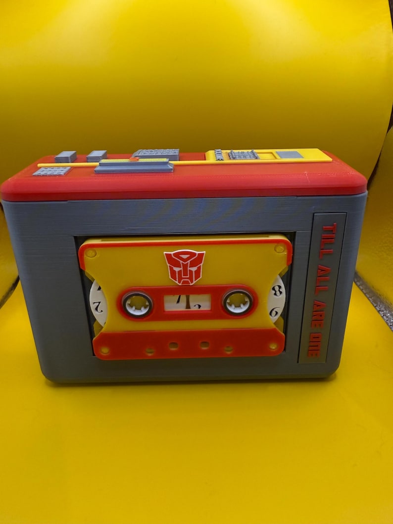 May include: A rectangular grey, red, and yellow plastic cassette player case featuring a yellow cassette tape with a red Autobot symbol in the centre. The side of the case displays the text TILL ALL ARE ONE in red letters against a grey background.