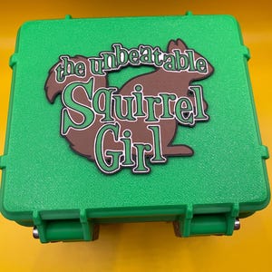 May include: A bright green square storage case with a textured surface and two front latches. Centered on the lid is a brown silhouette of a squirrel with the text "the unbeatable Squirrel Girl" printed in green with a black outline.