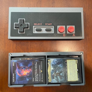 May include: A gray and black controller-shaped box with red buttons and the text "SELECT" and "START". Below is a gray card holder containing two stacks of Magic: The Gathering cards.