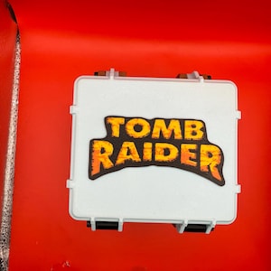 May include: A white rectangular case with black latches and hinges. The case has a sticker with the words "TOMB RAIDER" in orange and yellow. The background is a bright red.