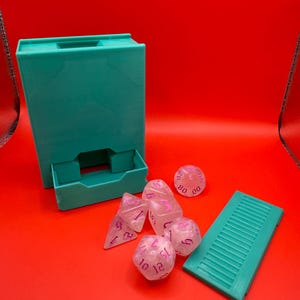 May include: A teal blue dice tower with a drawer and a matching teal blue dice tray. Six clear dice with pink numbers are scattered around the tower and tray.