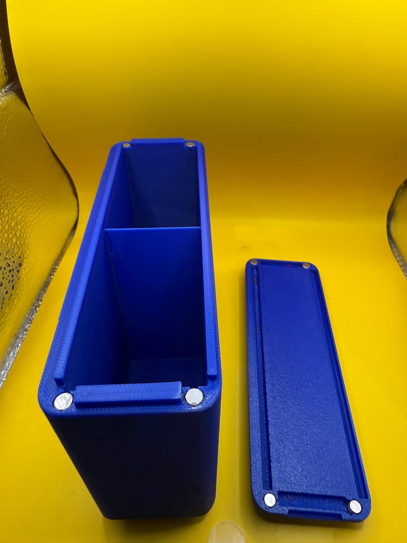 May include: A rectangular blue storage container with a divided interior and a matching detachable lid. The container features small circular silver magnets embedded at the corners of the opening and the underside of the lid for secure closure.