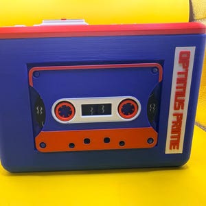 May include: A rectangular blue storage box featuring a red top edge and a prominent cassette tape design on the front. The cassette is blue, white, and orange with the text OPTIMUS PRIME printed vertically in red on a white rectangular label.