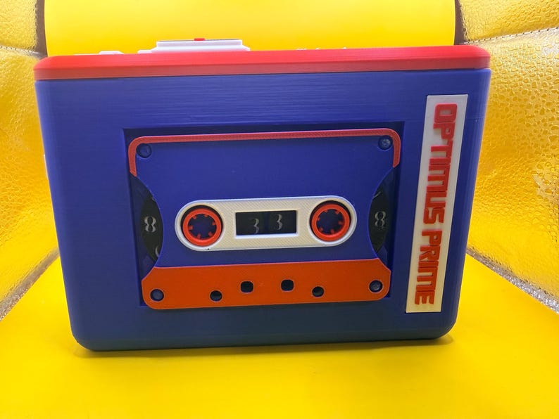 May include: A rectangular blue storage box featuring a red top edge and a prominent cassette tape design on the front. The cassette is blue, white, and orange with the text OPTIMUS PRIME printed vertically in red on a white rectangular label.