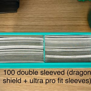 May include: A rectangular teal plastic storage tray containing two stacks of trading cards. The cards are protected by double sleeves, specifically Dragon Shield and Ultra Pro Fit brands. The text 100 double sleeved (dragon shield + ultra pro fit sleeves) is printed below the tray.
