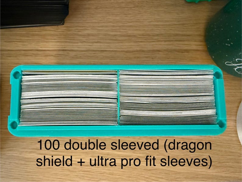 May include: A rectangular teal plastic storage tray containing two stacks of trading cards. The cards are protected by double sleeves, specifically Dragon Shield and Ultra Pro Fit brands. The text 100 double sleeved (dragon shield + ultra pro fit sleeves) is printed below the tray.