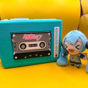 May include: A teal rectangular cassette player-shaped clock with a grey cassette tape insert featuring the text Hatsune Miku and Japanese characters. Next to it sits a small, plush, light blue-haired character doll with a smiling expression.