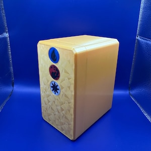 May include: A gold-colored, rectangular box with a textured surface. The box has three circular symbols on the front: a blue water drop, a red dragon, and a yellow sun.