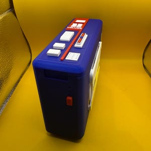 May include: A blue rectangular portable cassette player featuring a top control panel with white buttons, a red volume slider labelled "VOLUME", and a small red side button. The device has a textured plastic finish and a white side-mounted component.