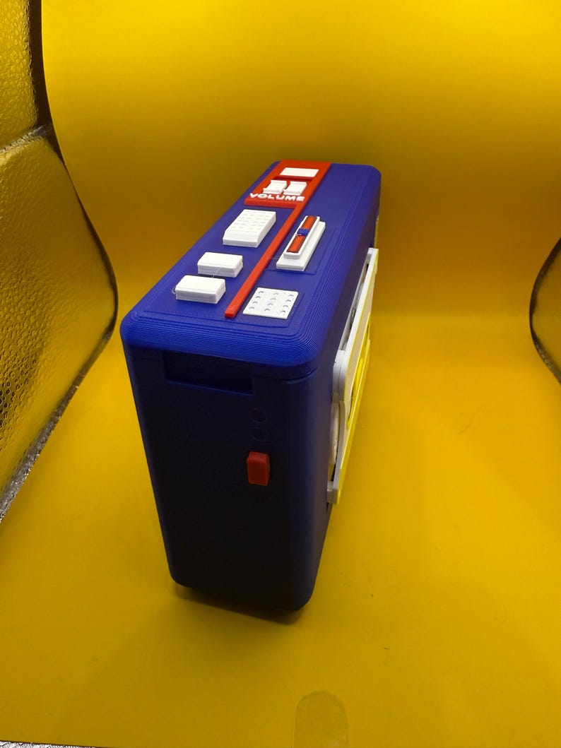 May include: A blue rectangular portable cassette player featuring a top control panel with white buttons, a red volume slider labelled "VOLUME", and a small red side button. The device has a textured plastic finish and a white side-mounted component.