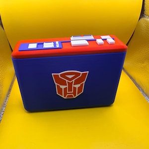 May include: A rectangular blue storage box featuring a prominent red and white Autobot logo on the front. The top lid is red with a blue panel containing white and blue buttons, switches, and the word VOLUME printed in white lettering.