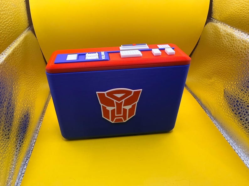 May include: A rectangular blue storage box featuring a prominent red and white Autobot logo on the front. The top lid is red with a blue panel containing white and blue buttons, switches, and the word VOLUME printed in white lettering.