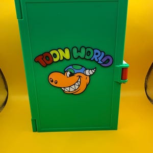 May include: A rectangular green case featuring the text TOON WORLD in colorful letters above a cartoon illustration of an orange creature with a blue and green spotted hat. The case has a small red latch on the right side and a smooth finish.