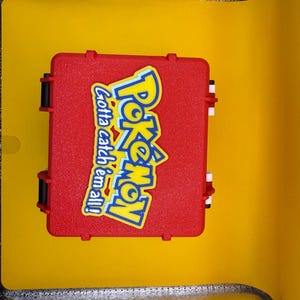 May include: A red plastic case with a Pokemon logo and the text "Gotta catch 'em all!" printed on it.