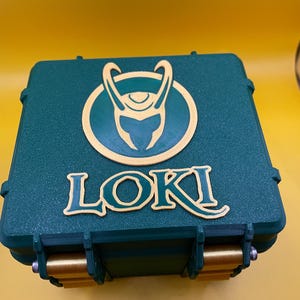 May include: A square, dark green storage case with a textured surface. The lid features a raised, light yellow circular emblem depicting a horned helmet above the word LOKI in a stylized font. Two gold-colored latches are visible at the front.