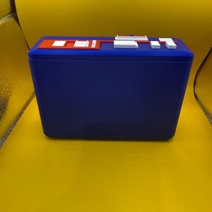 May include: A rectangular blue plastic case featuring a top panel with a red recessed section containing white buttons and a slider labelled VOLUME. The top surface also includes additional white rectangular buttons and a small blue switch.