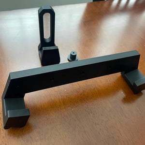 May include: A black plastic wall mount with two hooks and a small, separate black plastic stand. The wall mount has a rectangular shape with rounded edges and a raised lip on the top and bottom. The stand has a rectangular base and a cylindrical top.