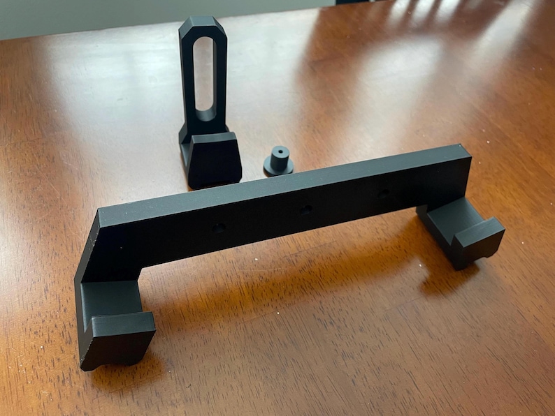 May include: A black plastic wall mount with two hooks and a small, separate black plastic stand. The wall mount has a rectangular shape with rounded edges and a raised lip on the top and bottom. The stand has a rectangular base and a cylindrical top.