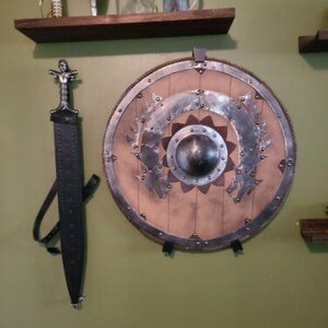 May include: A brown wooden shelf with a sword and a shield hanging on the wall behind it. The sword has a black leather scabbard and a silver handle. The shield is round and has a brown leather backing with a silver metal rim. The shield has a decorative design of a flower with a silver metal center.