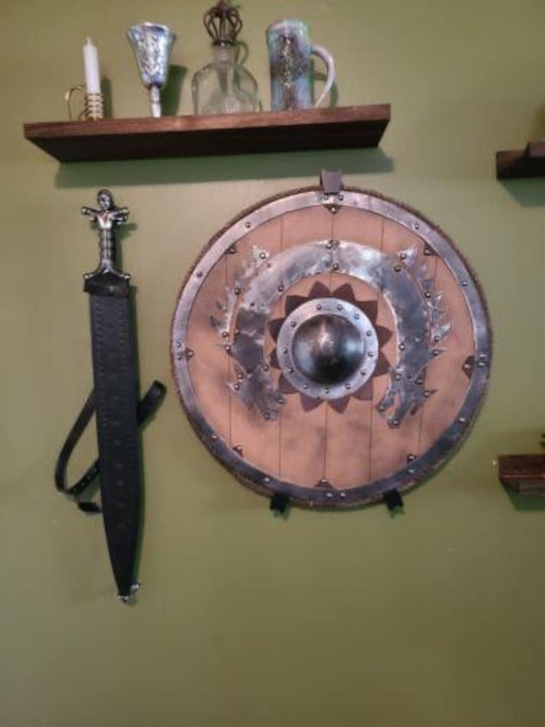 May include: A brown wooden shelf with a sword and a shield hanging on the wall behind it. The sword has a black leather scabbard and a silver handle. The shield is round and has a brown leather backing with a silver metal rim. The shield has a decorative design of a flower with a silver metal center.