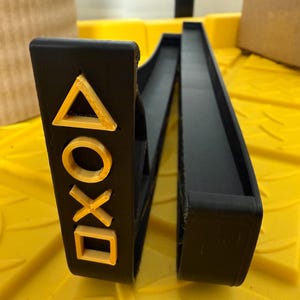 May include: Black plastic stand with a cutout design of the PlayStation controller symbols in gold. The stand is open and ready to hold a controller.