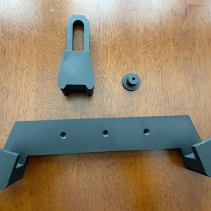 May include: A black plastic wall mount with three holes and two angled brackets. The mount is designed to hold a device or object.