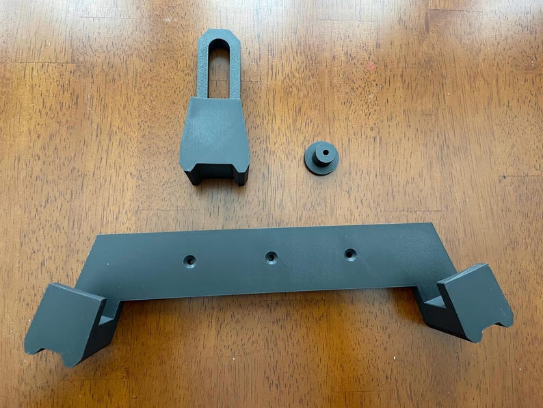 May include: A black plastic wall mount with three holes and two angled brackets. The mount is designed to hold a device or object.