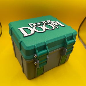 Op de afbeelding: Een vierkante opbergkoffer met harde schaal, een groen deksel en een grijze basis. Op het deksel staat de tekst DOCTOR DOOM in verhoogde witte letters met een zwarte omlijning. De koffer is voorzien van groene sluitingen met zilveren schroeven.