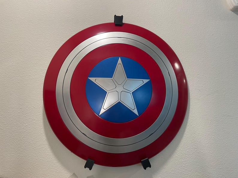 May include: A red, white, and blue Captain America shield with a silver star in the center. The shield is mounted on a white wall with black brackets.