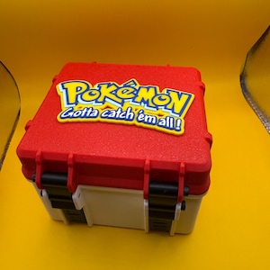 May include: A red and white plastic storage box with a black latch. The lid has a yellow Pokemon logo with the text "Gotta catch 'em all!" in blue.