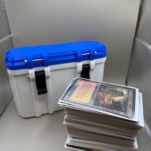 May include: A white and blue plastic storage box with a black handle and a stack of white trading cards with a colourful card on top. The top card has a picture of a large stone structure with a red and orange sky in the background. The text on the card reads "City of Brass".