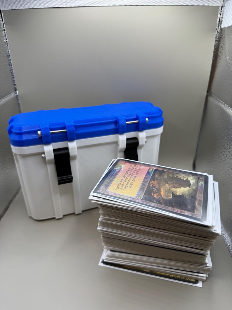 May include: A white and blue plastic storage box with a black handle and a stack of white trading cards with a colourful card on top. The top card has a picture of a large stone structure with a red and orange sky in the background. The text on the card reads "City of Brass".