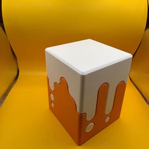 Puede incluir: Una caja cuadrada de dos tonos con la parte superior blanca y la base naranja. La sección naranja presenta un diseño de goteo con acentos redondeados. La caja está sobre un fondo amarillo brillante.