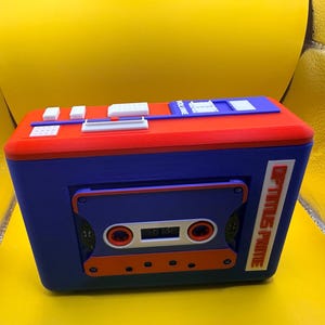 May include: A rectangular blue and red plastic cassette player model featuring a central cassette tape insert, white control buttons on the top surface, and the text OPTIMUS PRIME printed vertically in orange on the right side of the front panel.