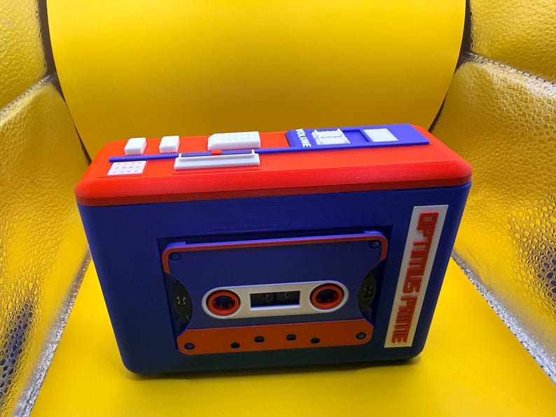 May include: A rectangular blue and red plastic cassette player model featuring a central cassette tape insert, white control buttons on the top surface, and the text OPTIMUS PRIME printed vertically in orange on the right side of the front panel.
