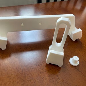 May include: White 3D printed plastic parts for a phone stand. The parts include a base, a vertical support, and a horizontal bar.