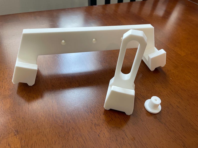 May include: White 3D printed plastic parts for a phone stand. The parts include a base, a vertical support, and a horizontal bar.