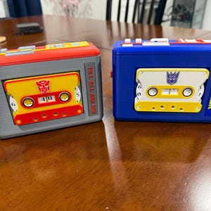 May include: Two rectangular plastic cassette player cases. The left case is grey and red with a yellow cassette featuring a red Autobot symbol and the text TILL ALL ARE ONE. The right case is blue with a white and yellow cassette featuring a purple Decepticon symbol and the text SUPERIOR.