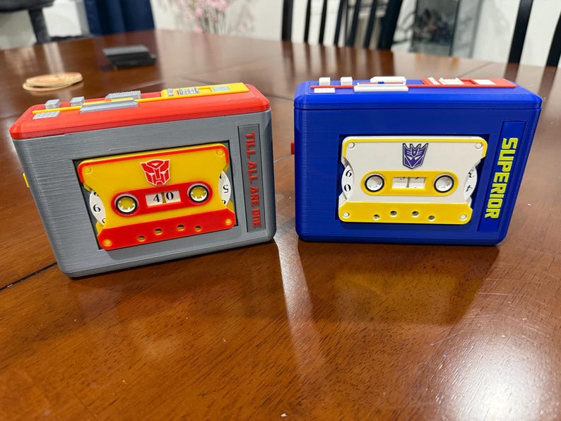 May include: Two rectangular plastic cassette player cases. The left case is grey and red with a yellow cassette featuring a red Autobot symbol and the text TILL ALL ARE ONE. The right case is blue with a white and yellow cassette featuring a purple Decepticon symbol and the text SUPERIOR.