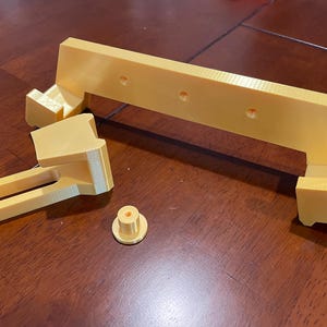 May include: Four pieces of a 3D printed, gold-colored plastic object. The pieces include a long, flat piece with holes, a smaller piece with a curved shape, a long, rectangular piece with a handle, and a small, round piece.