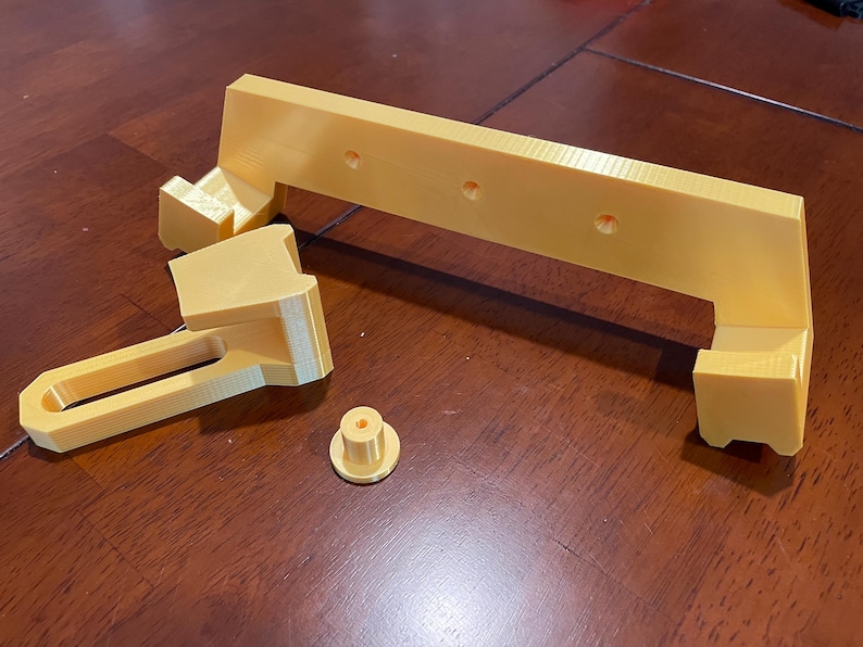 May include: Four pieces of a 3D printed, gold-colored plastic object. The pieces include a long, flat piece with holes, a smaller piece with a curved shape, a long, rectangular piece with a handle, and a small, round piece.