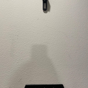 May include: Black wall mount for a video game console. The mount has two parts, a top part that is attached to the wall and a bottom part that is attached to the console. The bottom part has three screws.