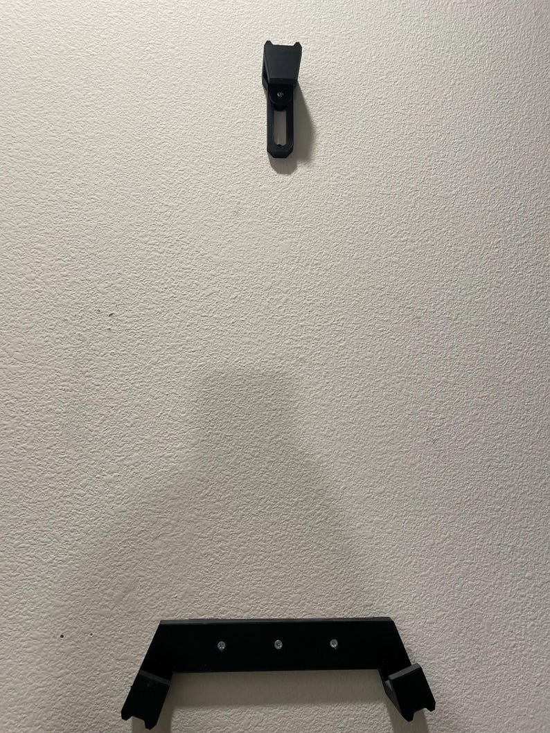 May include: Black wall mount for a video game console. The mount has two parts, a top part that is attached to the wall and a bottom part that is attached to the console. The bottom part has three screws.