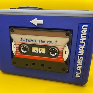 May include: A blue rectangular cassette player replica featuring a white arrow pointing left and the text PLANES WALKMAN on the right side. A cassette tape labeled AWESOME MIX VOL. 1 with red and white stripes is centered in the front display window.